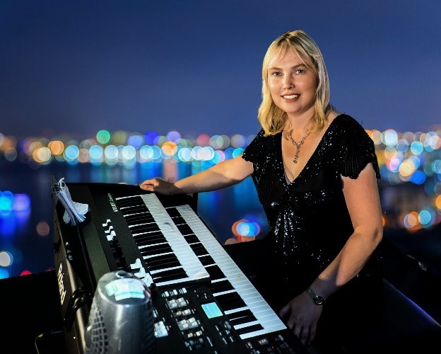 Image of Elizabeth Harrison sitting at a black organ with a row of blurred coloured lights behind her as through a window